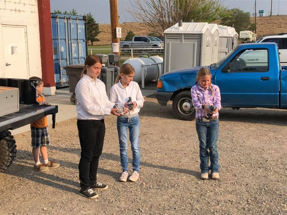 Three young girls are standing in front of a blue truck holding remote controls.