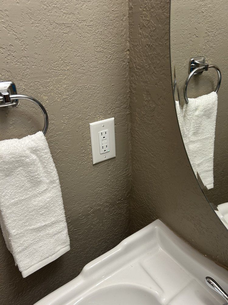 Bathroom sink corner with towel ring, white hand towel, mirror reflection, and wall outlet; countertop at bottom.