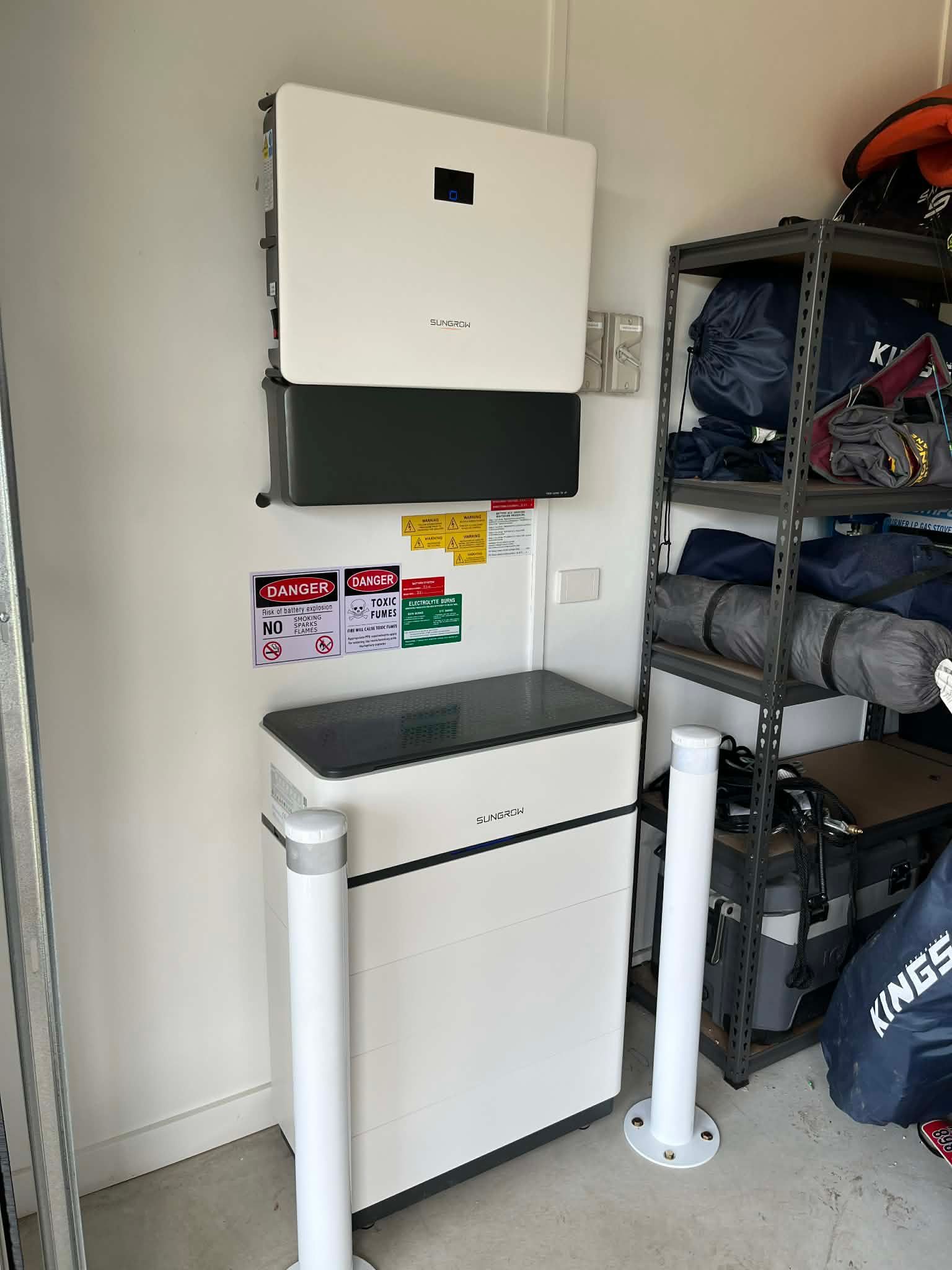 White Solar Inverter And Battery On A Wall, With Safety Signs And A Metal Shelving Unit Nearby — J T Walker Solar & Electrical In Taree, NSW