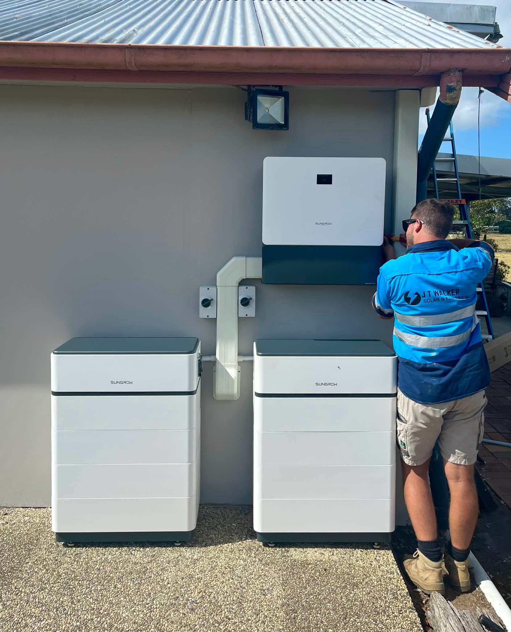 Electrician Installing Solar Batteries On A Building. White Boxes, Gray Wall, Blue And White Shirt — J T Walker Solar & Electrical In Taree, NSW