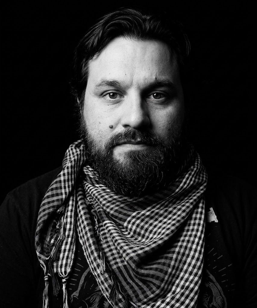 Black-and-white portrait of a bearded person wearing a patterned scarf, facing the camera with a neutral expression