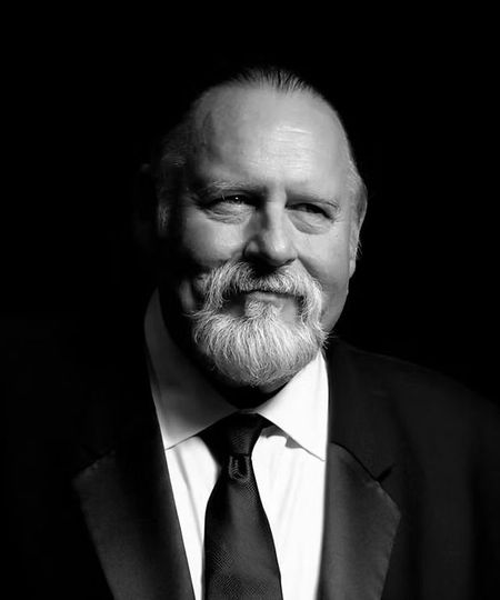 Black-and-white studio portrait of a bearded man in a suit and tie, faintly smiling against a dark background