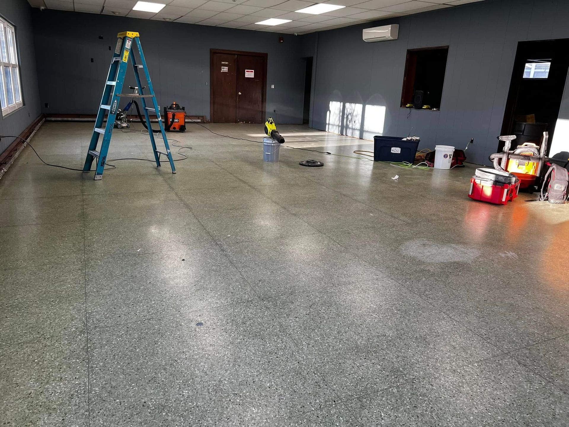 Empty room with gray floor, blue ladder, and cleaning supplies near the wall under bright lights