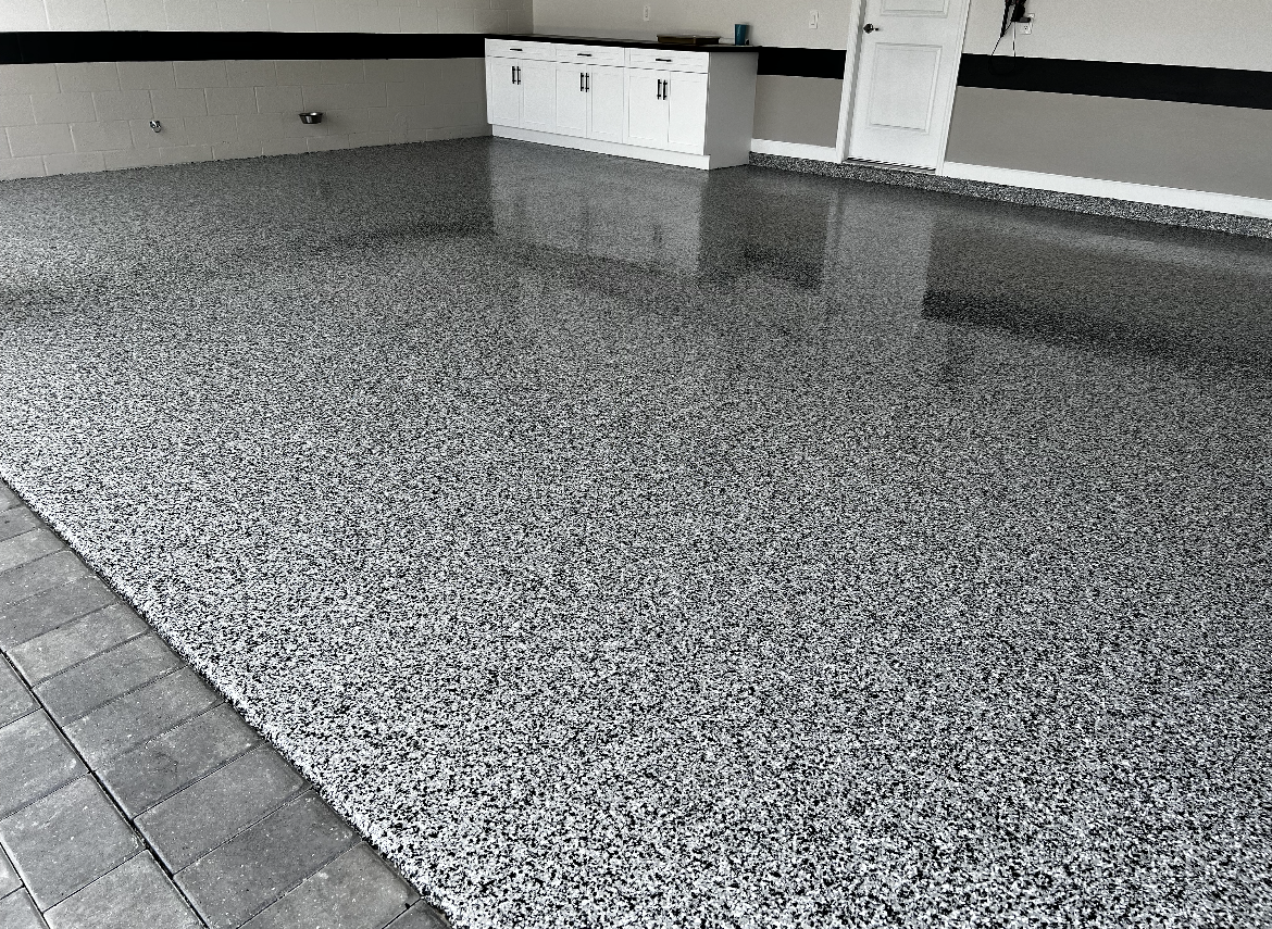 Speckled epoxy floor in a garage or utility area with a tiled edge and white storage boxes near the wall