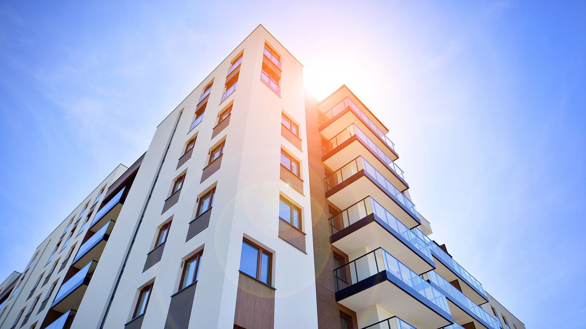 A tall, modern apartment building with balconies, facing the bright blue sky and sunlight.