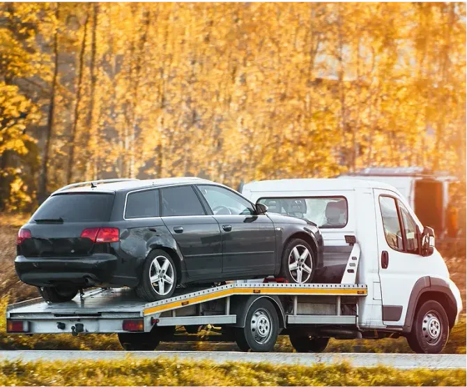 Black station wagon loaded on a flatbed tow truck beside a white van on an autumn roadside.