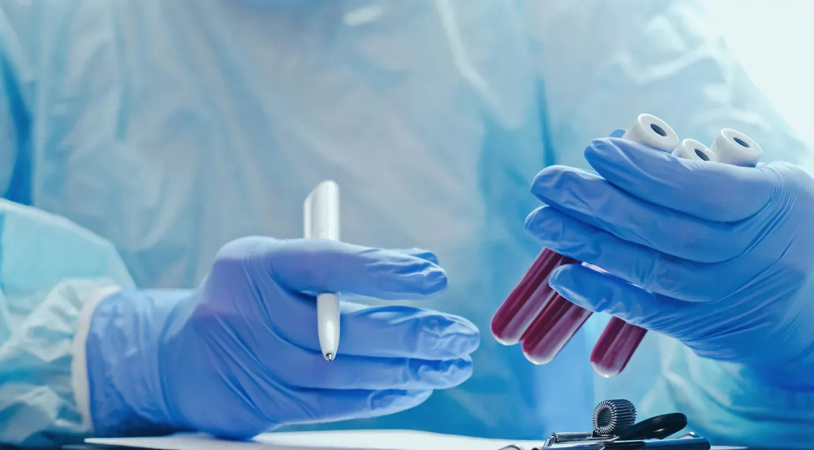 Person in blue medical gloves holding blood vials and a pen.