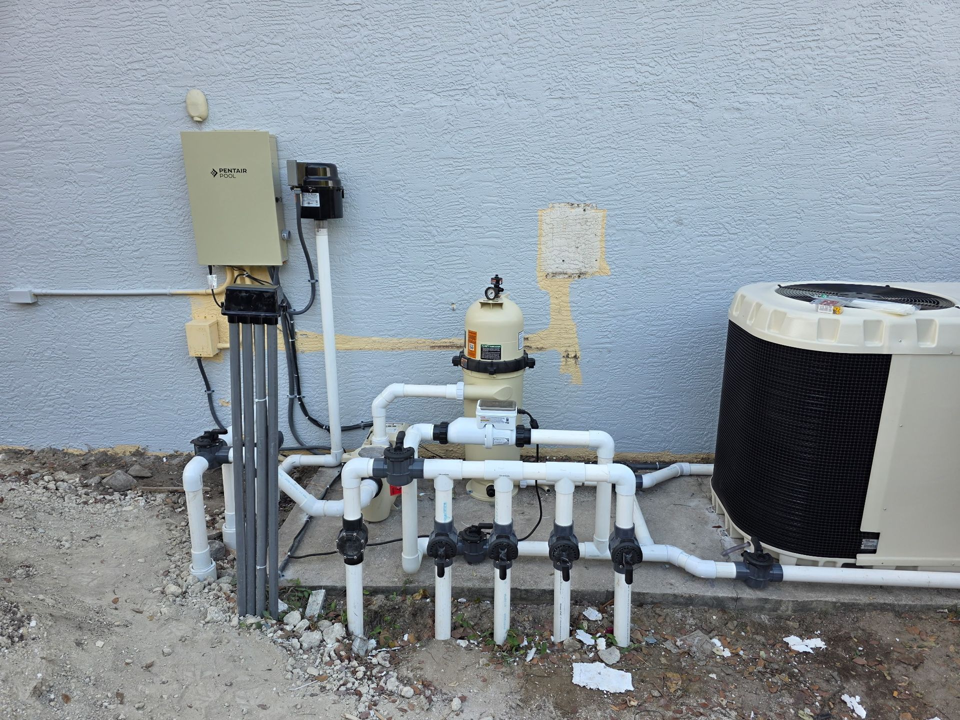 Outdoor HVAC and plumbing equipment mounted against a stucco wall, with pipes, valves, and a large fan unit.