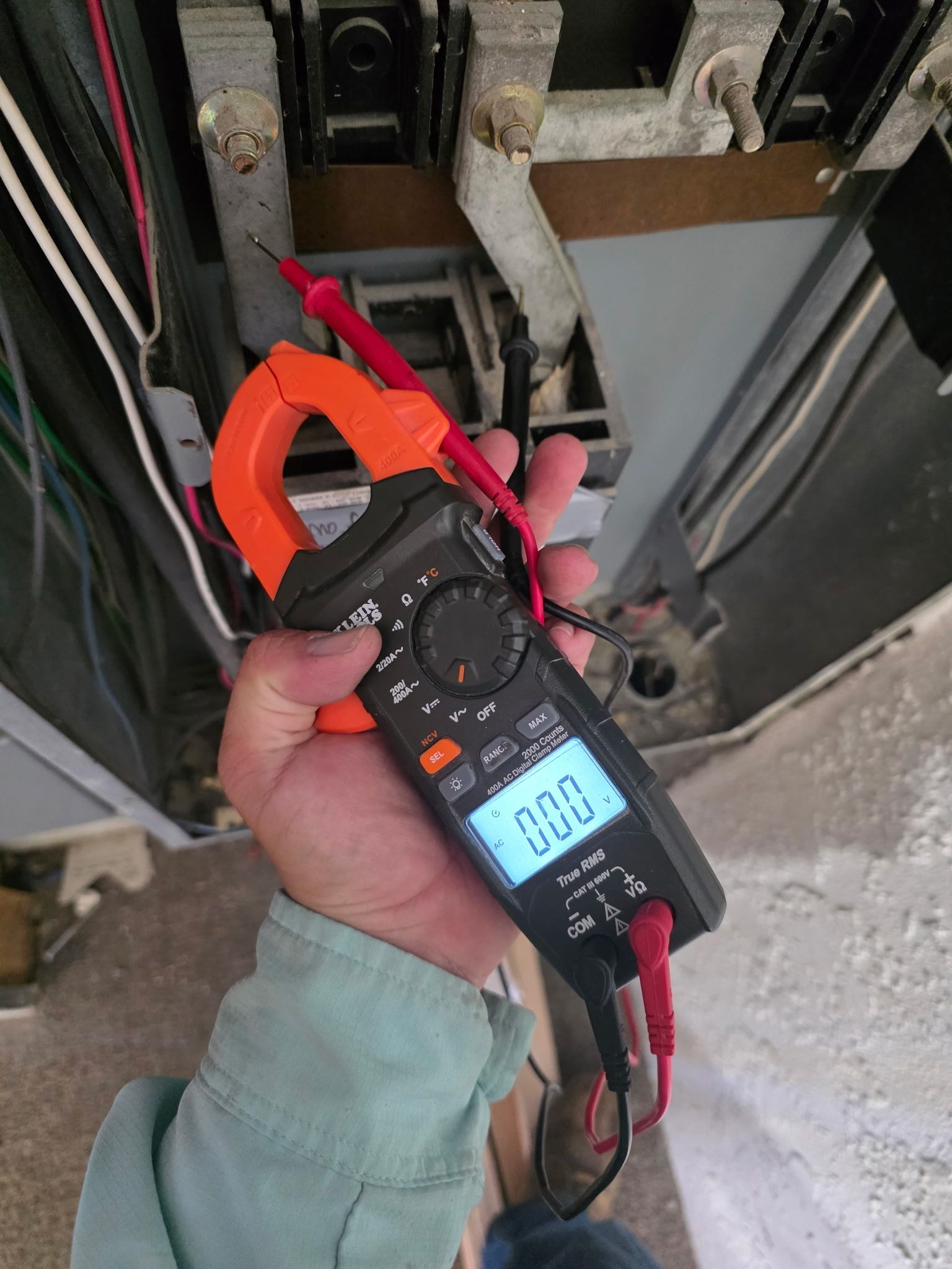 Hand holding a digital clamp meter with orange jaws near exposed electrical wiring and conduit in a utility area