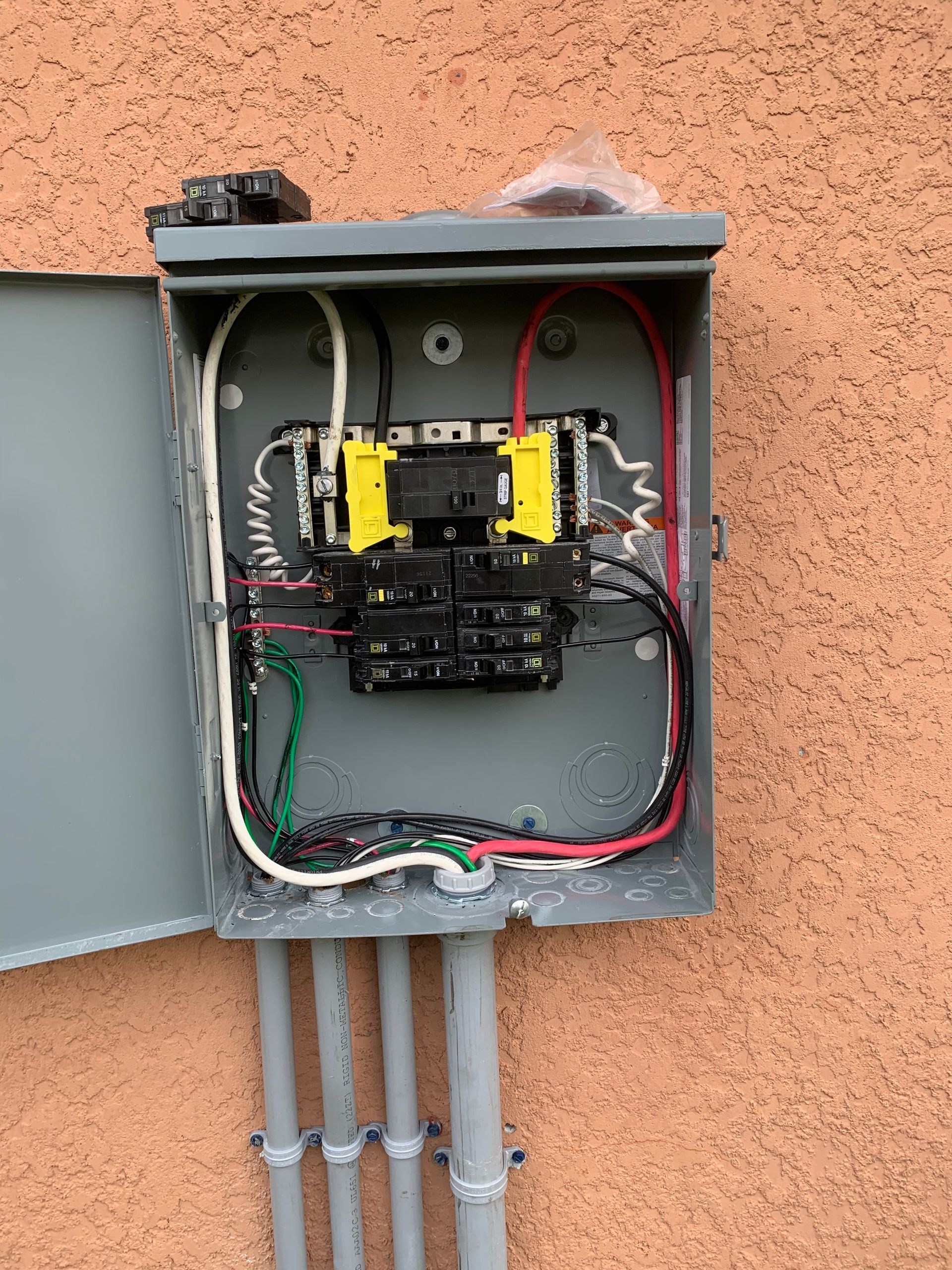 Open electrical panel with exposed wiring, breakers, and gray conduit on an exterior wall