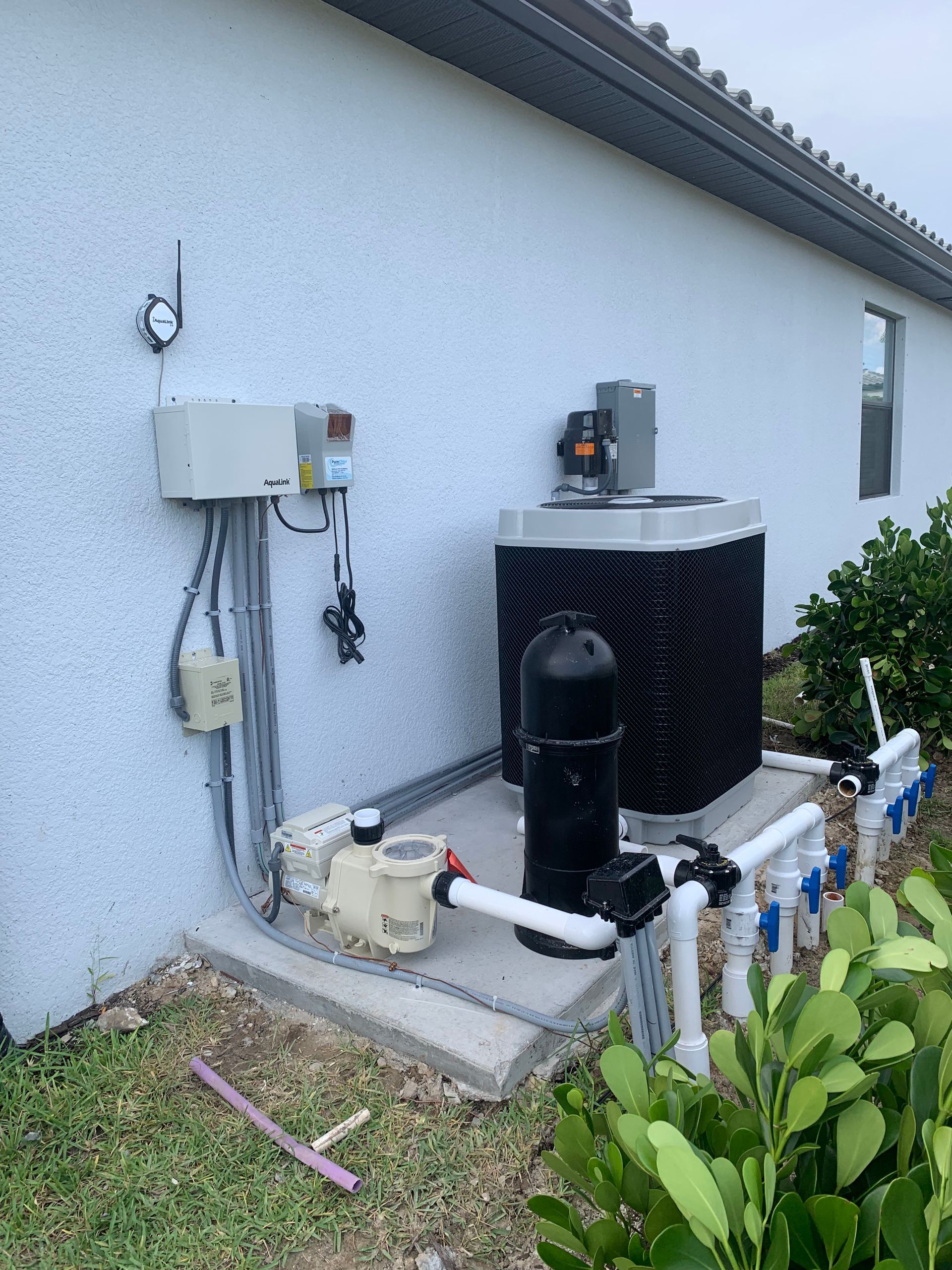 Outdoor air conditioner and heat pump units beside a white house wall with pipes and electrical boxes.