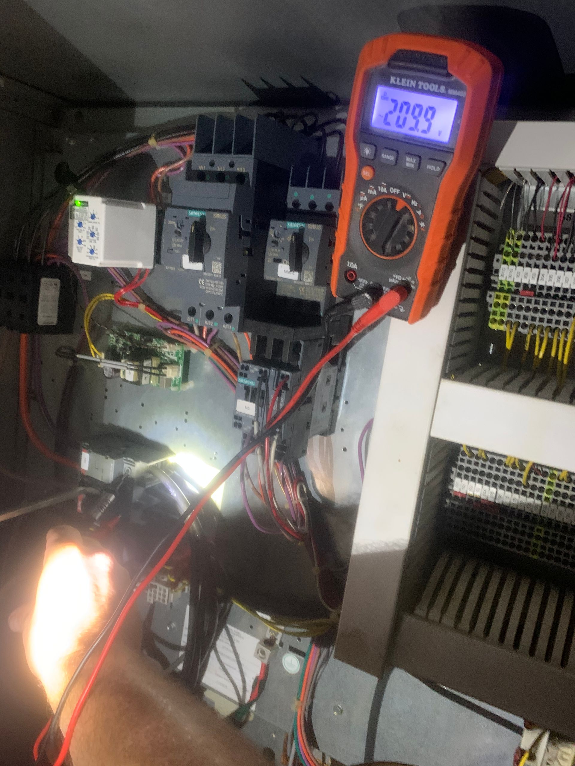 Orange digital multimeter clipped to an electrical panel, measuring a circuit with wires and terminal blocks.