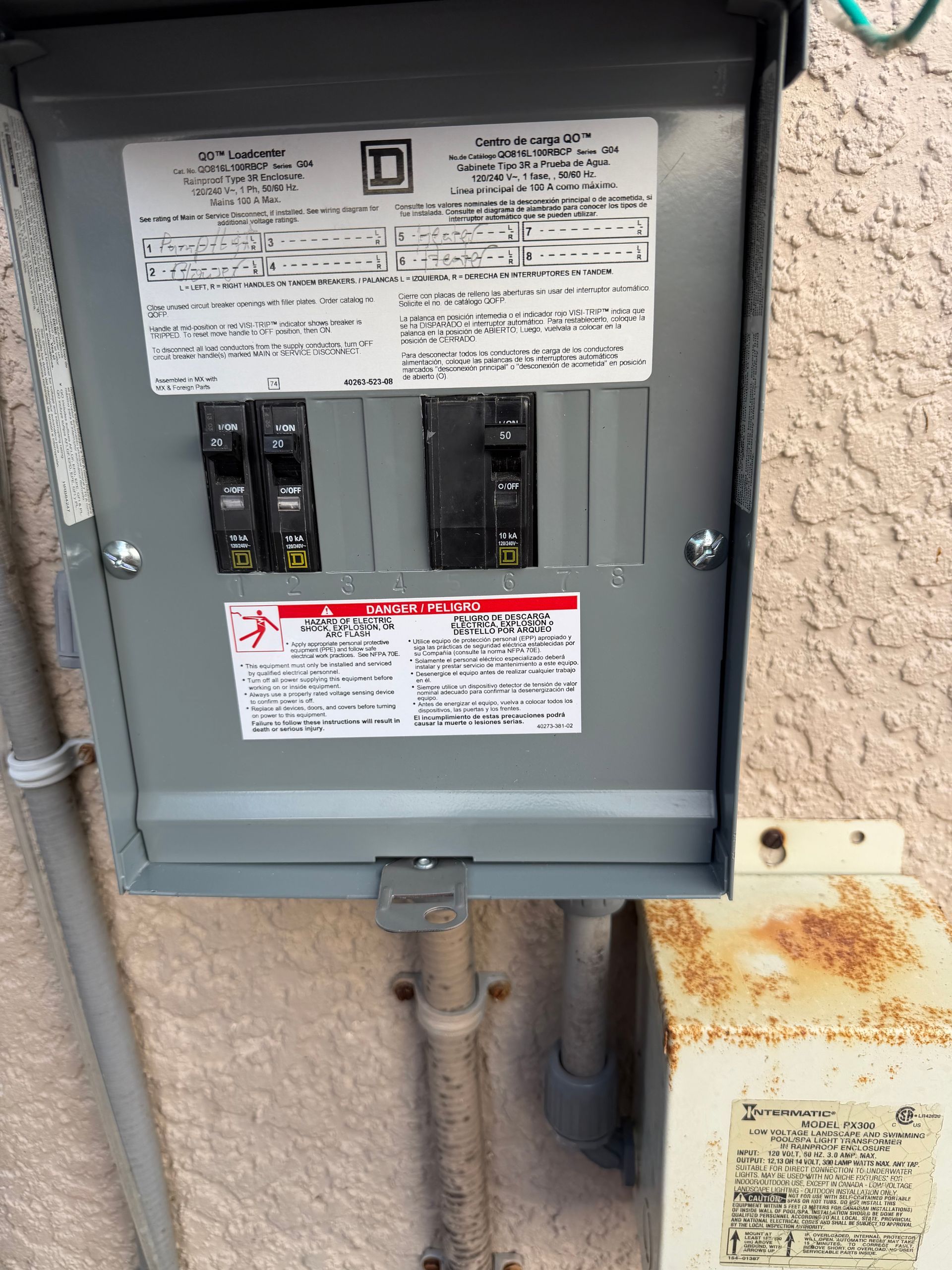 Open electrical breaker panel mounted on an exterior wall with rusted conduit below.