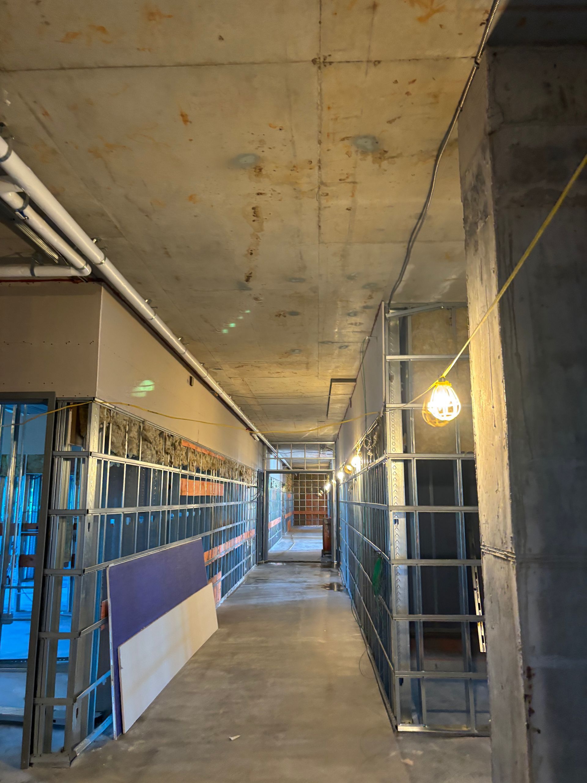 Narrow unfinished corridor with concrete walls, metal scaffolding, and work lights along one side.