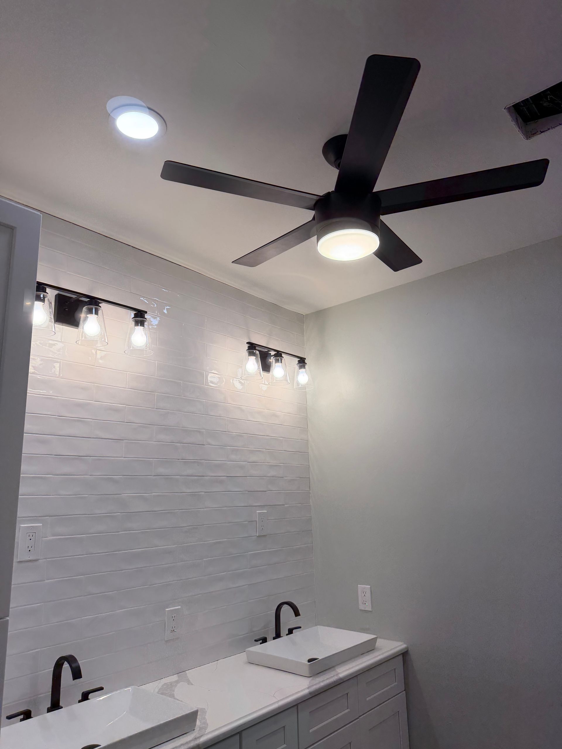 Modern bathroom with double vanity, white subway tile, black ceiling fan, and vanity lights