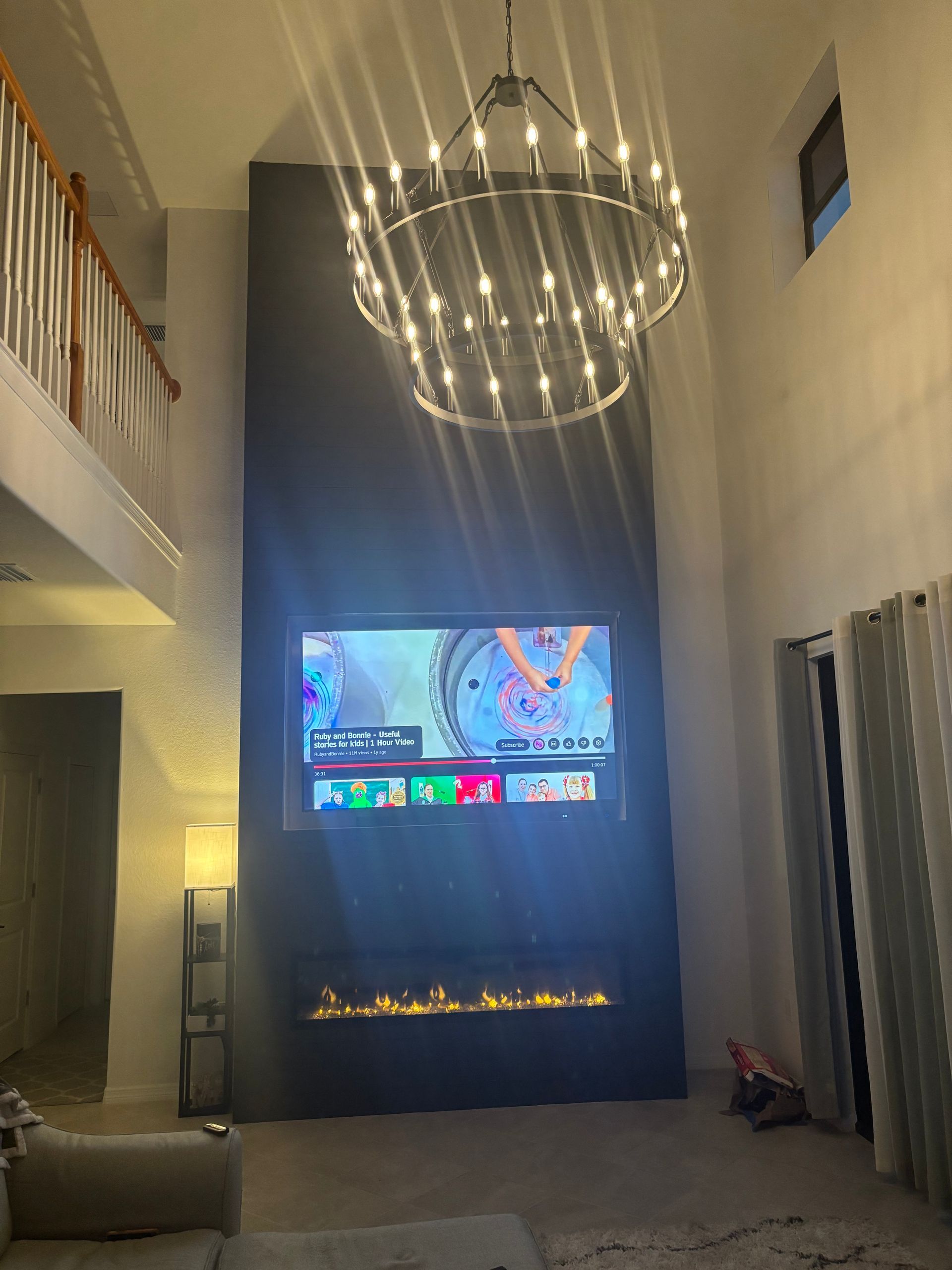 Modern living room with high ceiling, chandelier, wall-mounted TV, and linear fireplace behind dark curtains