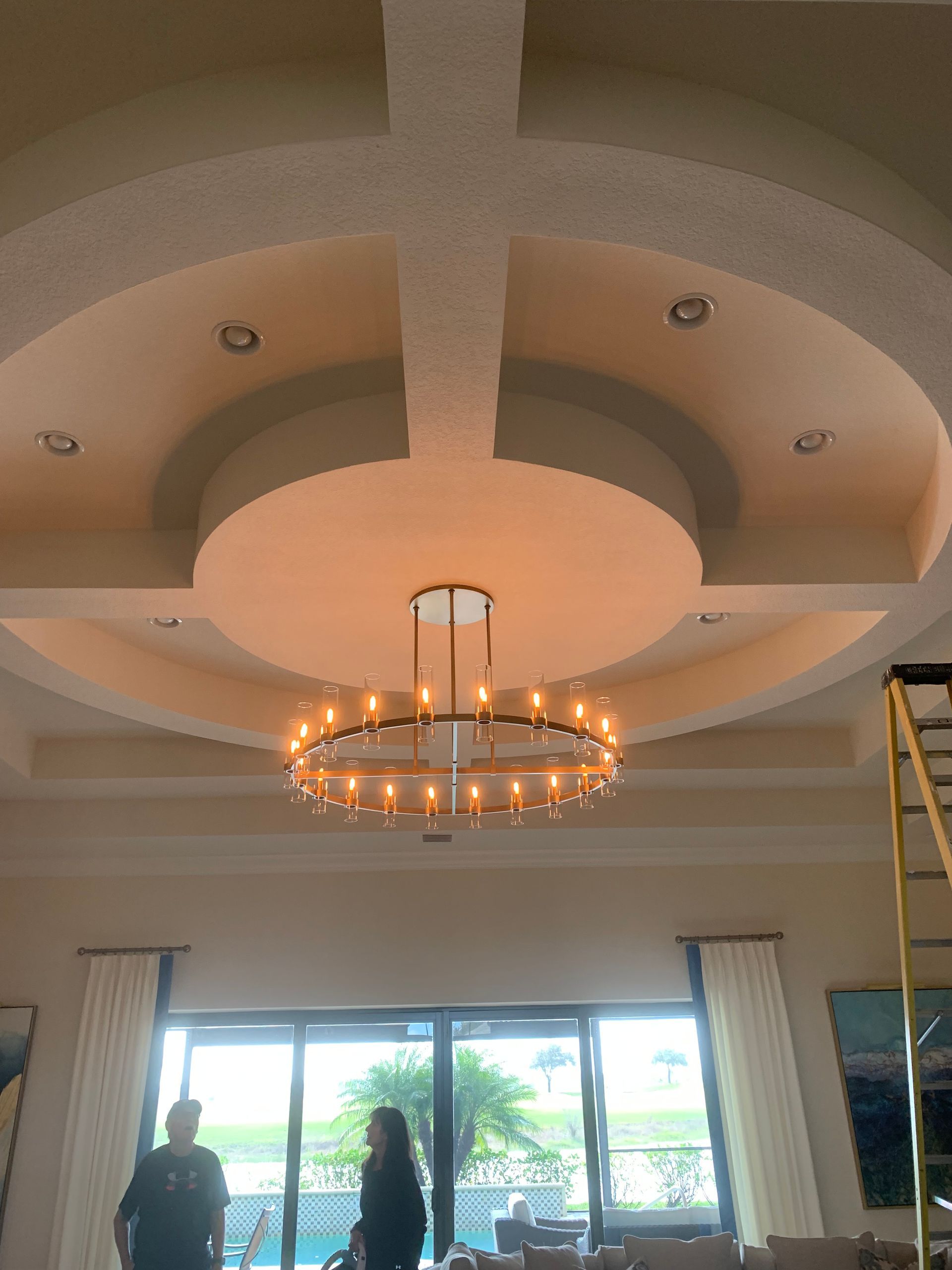 Elegant living room ceiling with chandelier, recessed lights, and large windows letting in daylight.