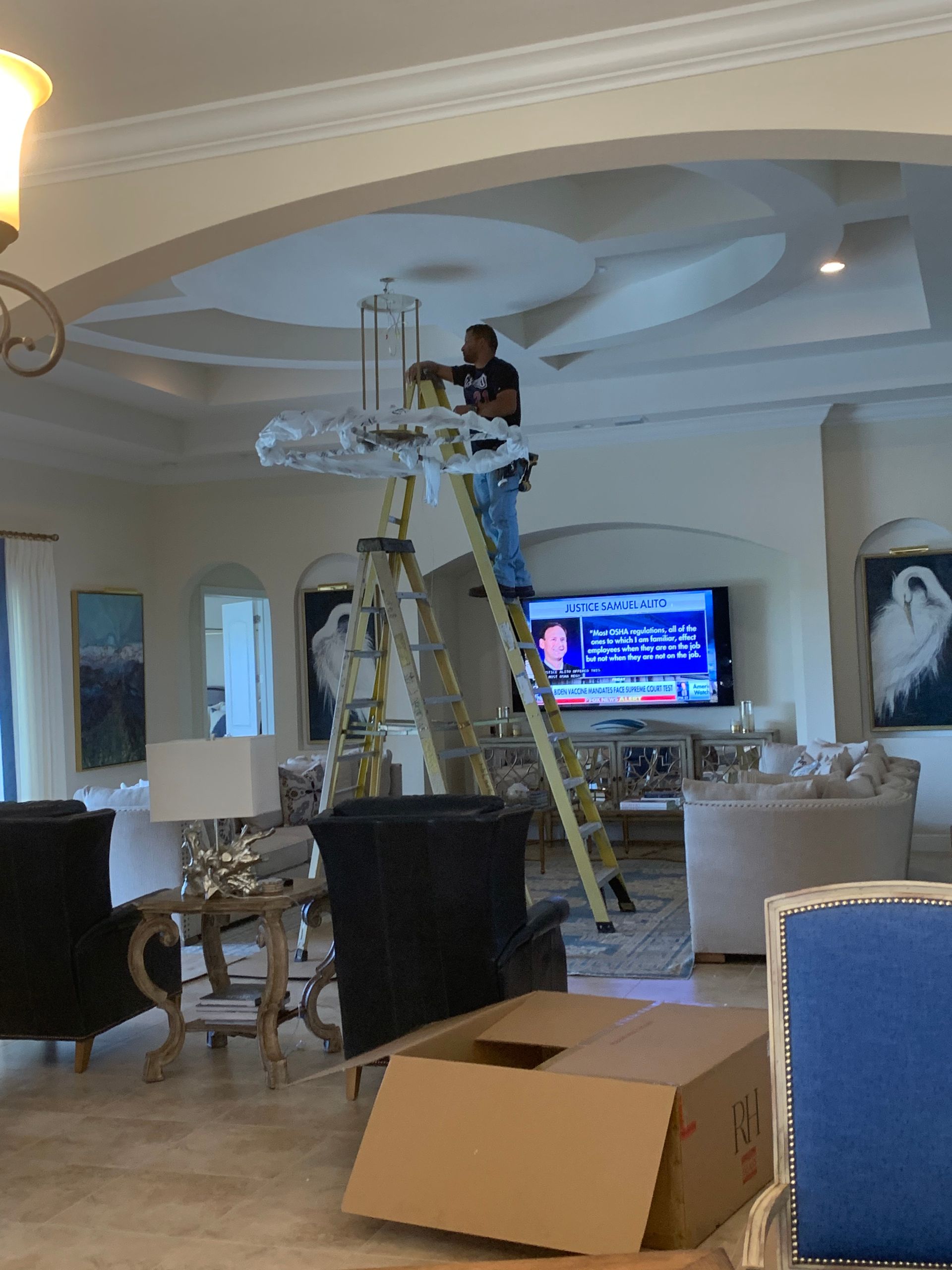 Person standing on a ladder fixing a ceiling light in a living room, with tools and an open box below.