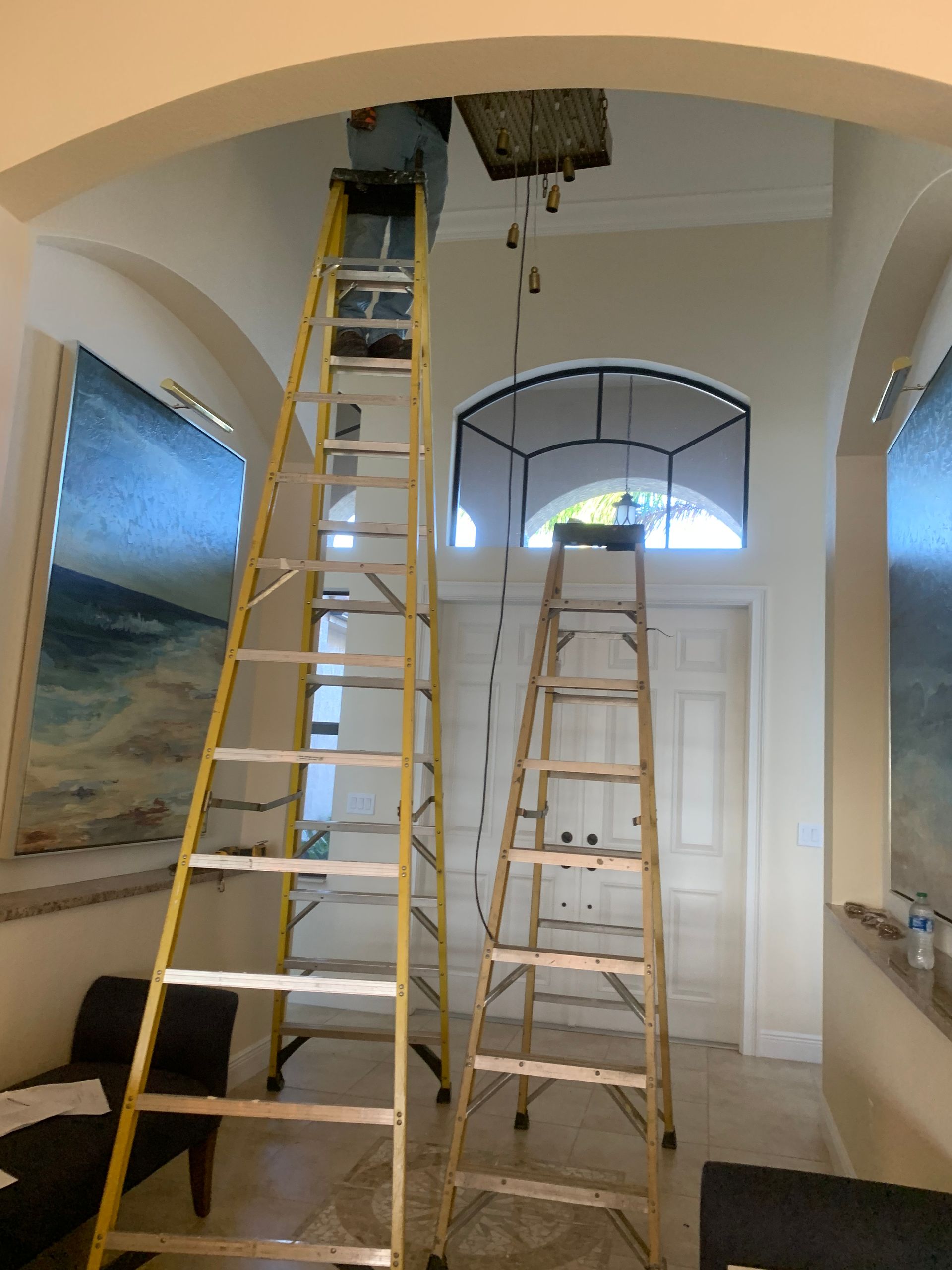 Two ladders in a hallway beneath a ceiling opening, with a wall mural and arched doorway nearby.