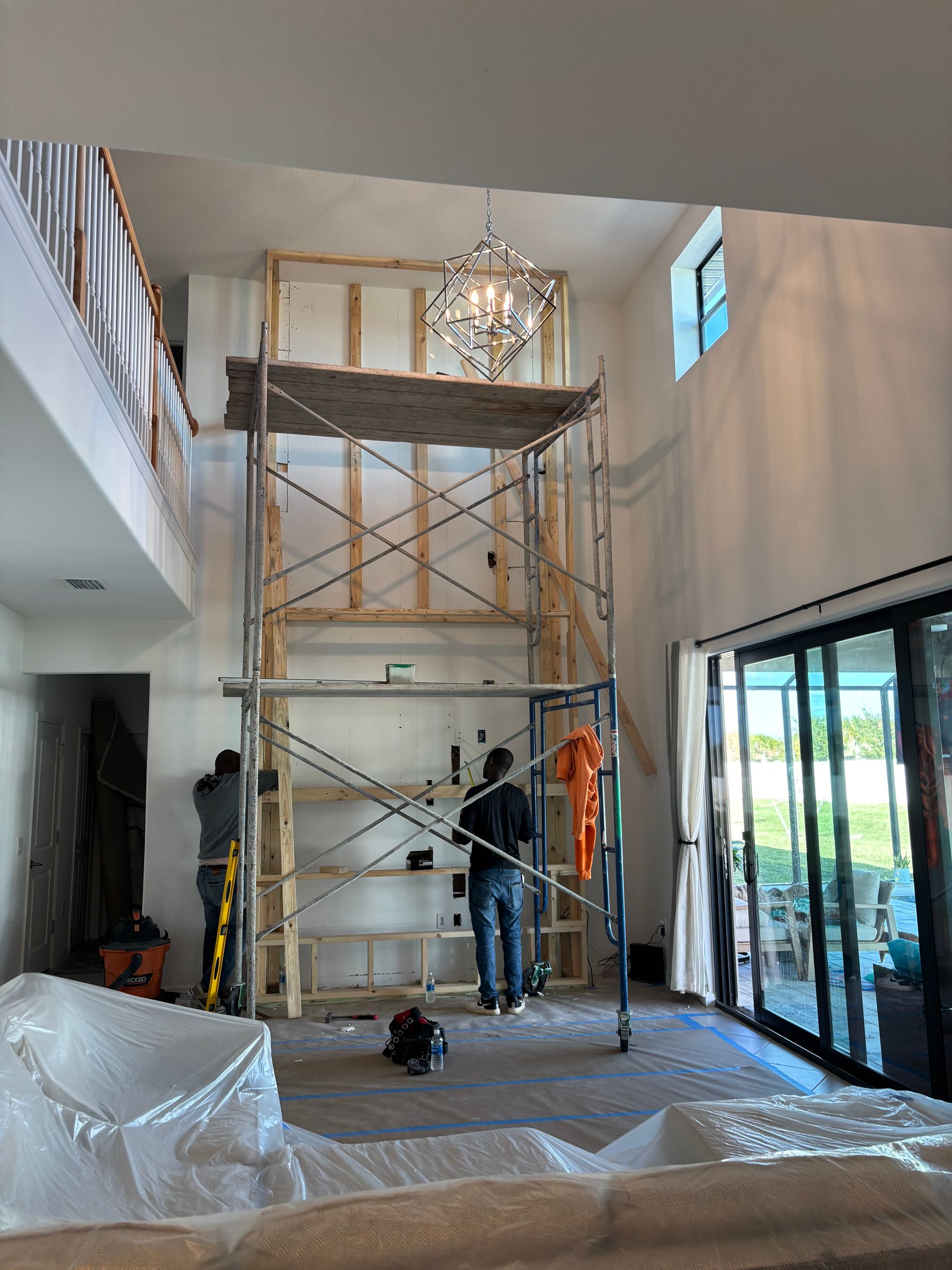 Workers on a scaffold in a high-ceilinged living room with plastic-covered furniture and large windows.