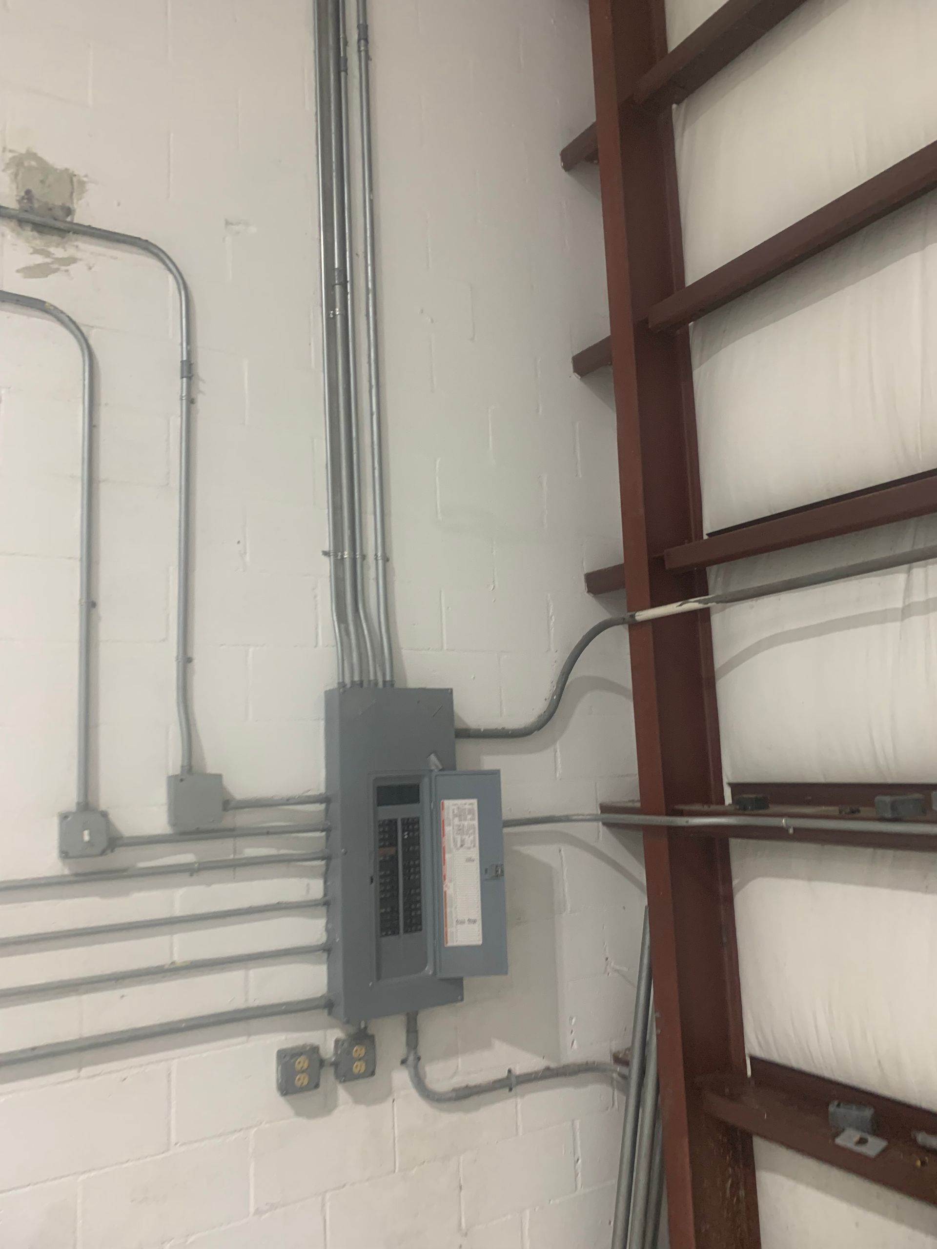 Electrical panel and conduit on a white cinderblock wall beside brown shelving.