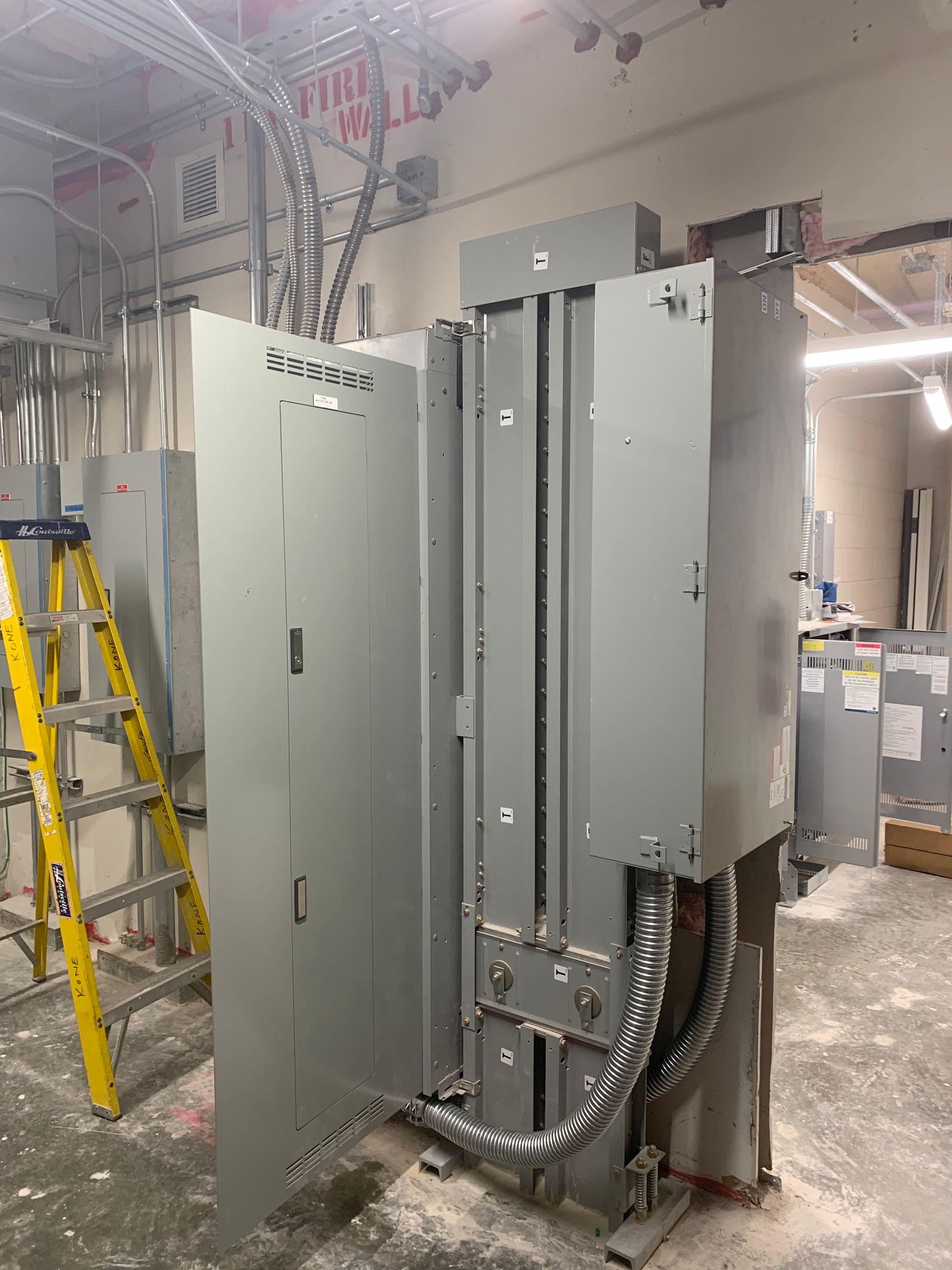 Electrical panels and conduit in an unfinished industrial room, with a yellow ladder at left.