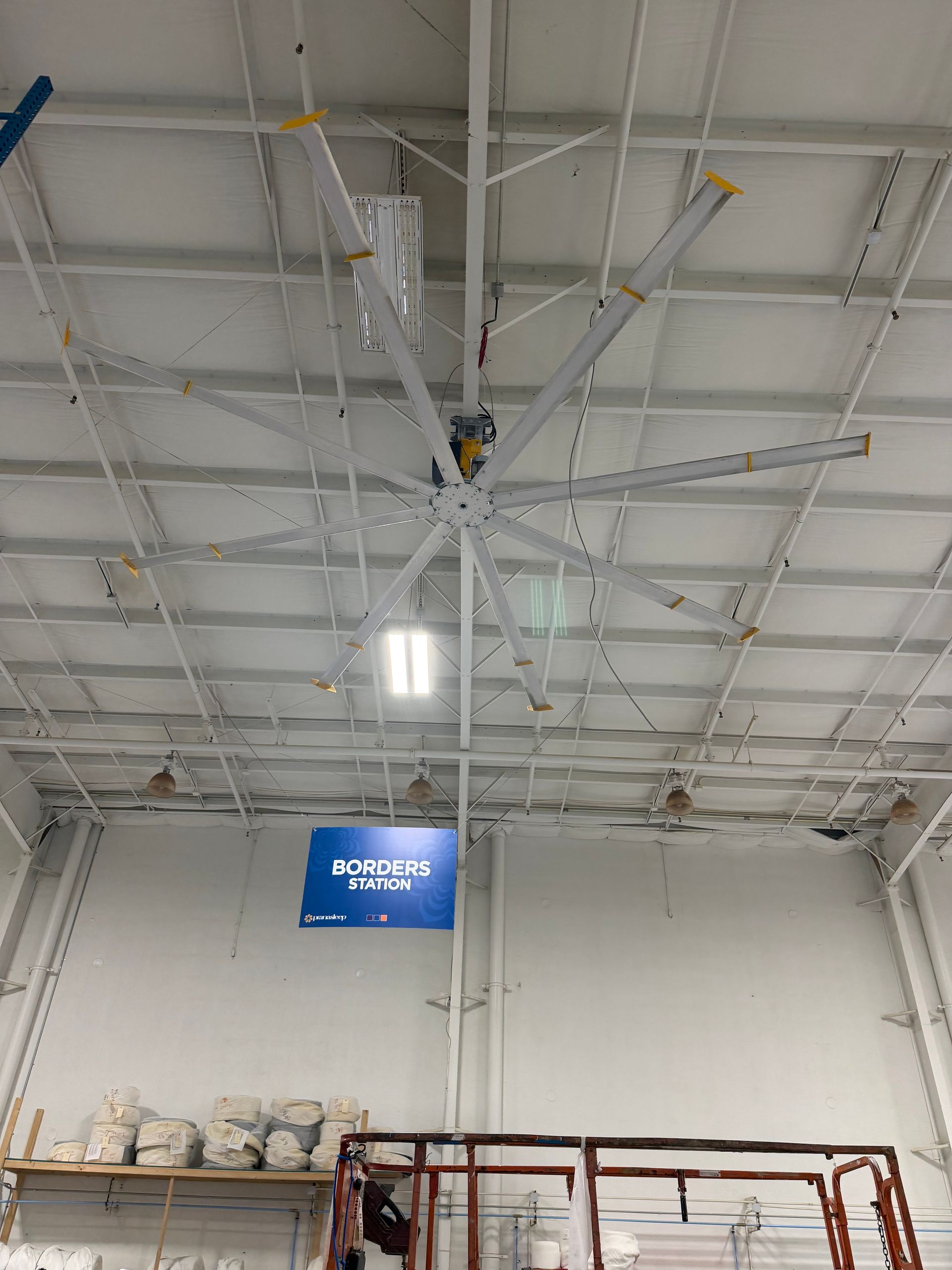 Warehouse ceiling with suspended equipment, bright lights, and a blue sign above shelves and a red scaffold.