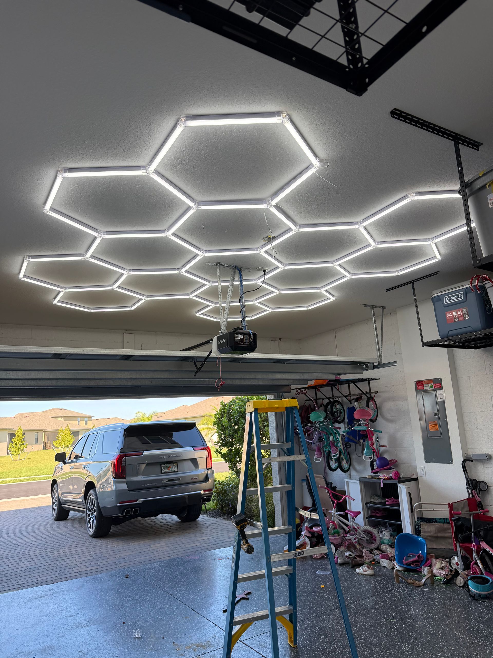 Garage with hexagon LED ceiling lights, parked SUV, ladder, and tools by open driveway.