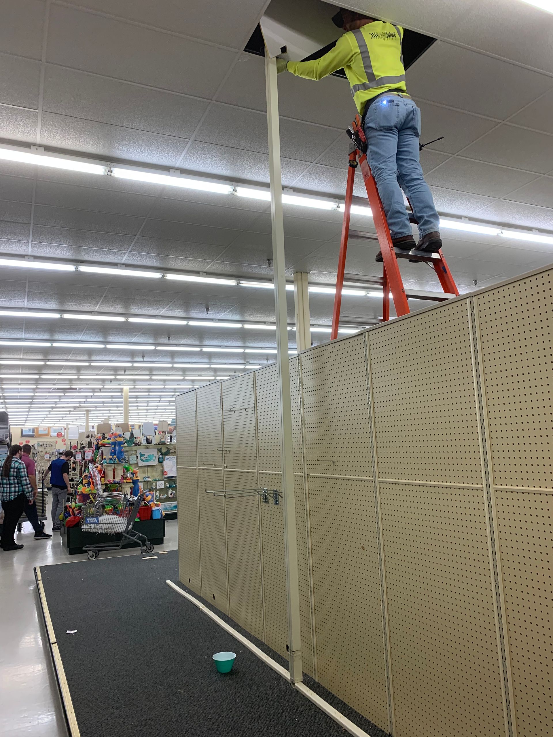 Worker on a red ladder installing a tall hanging sign in a store aisle.