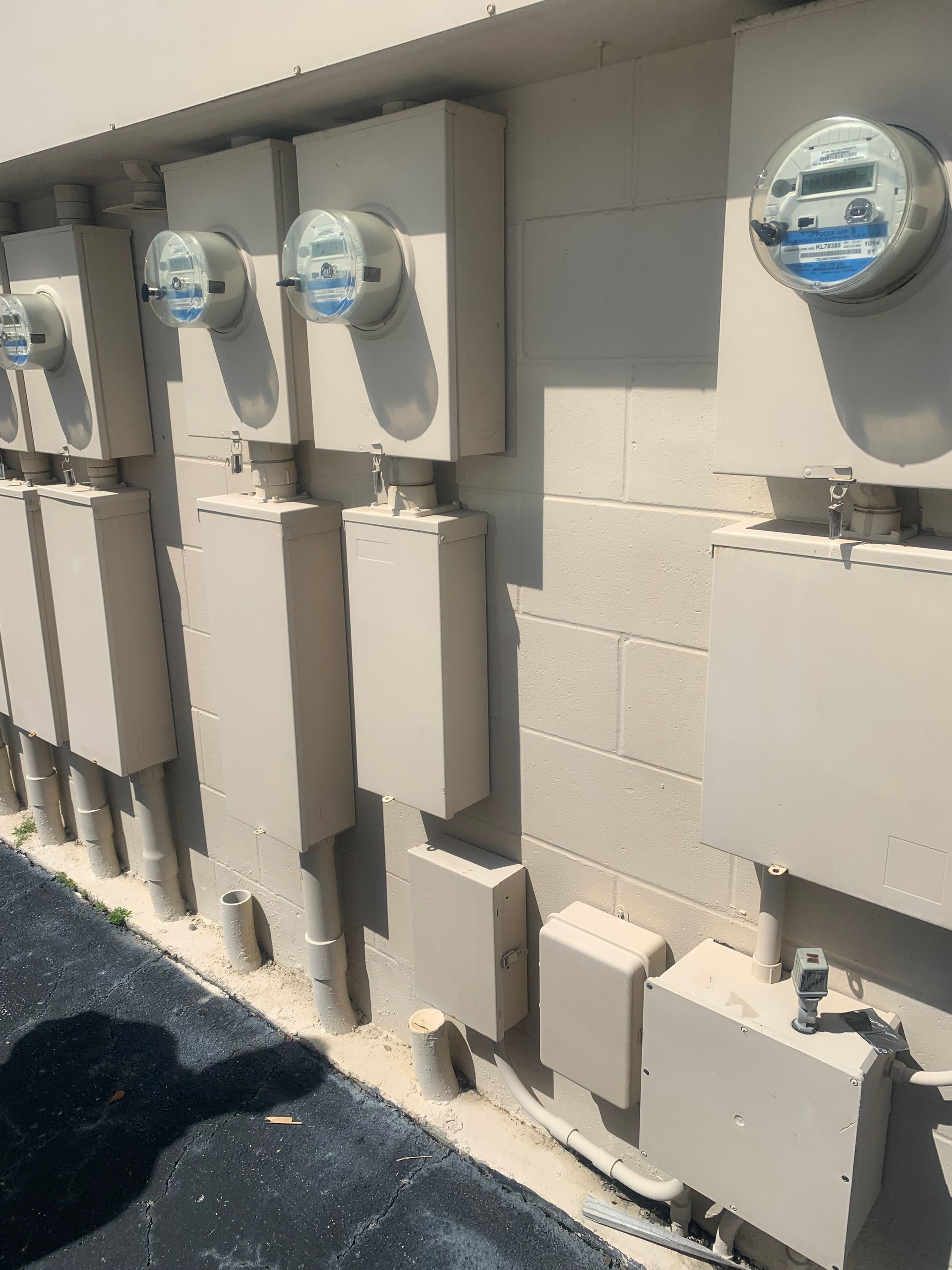 Row of electric utility meters and gray boxes mounted on a beige exterior wall in sunlight