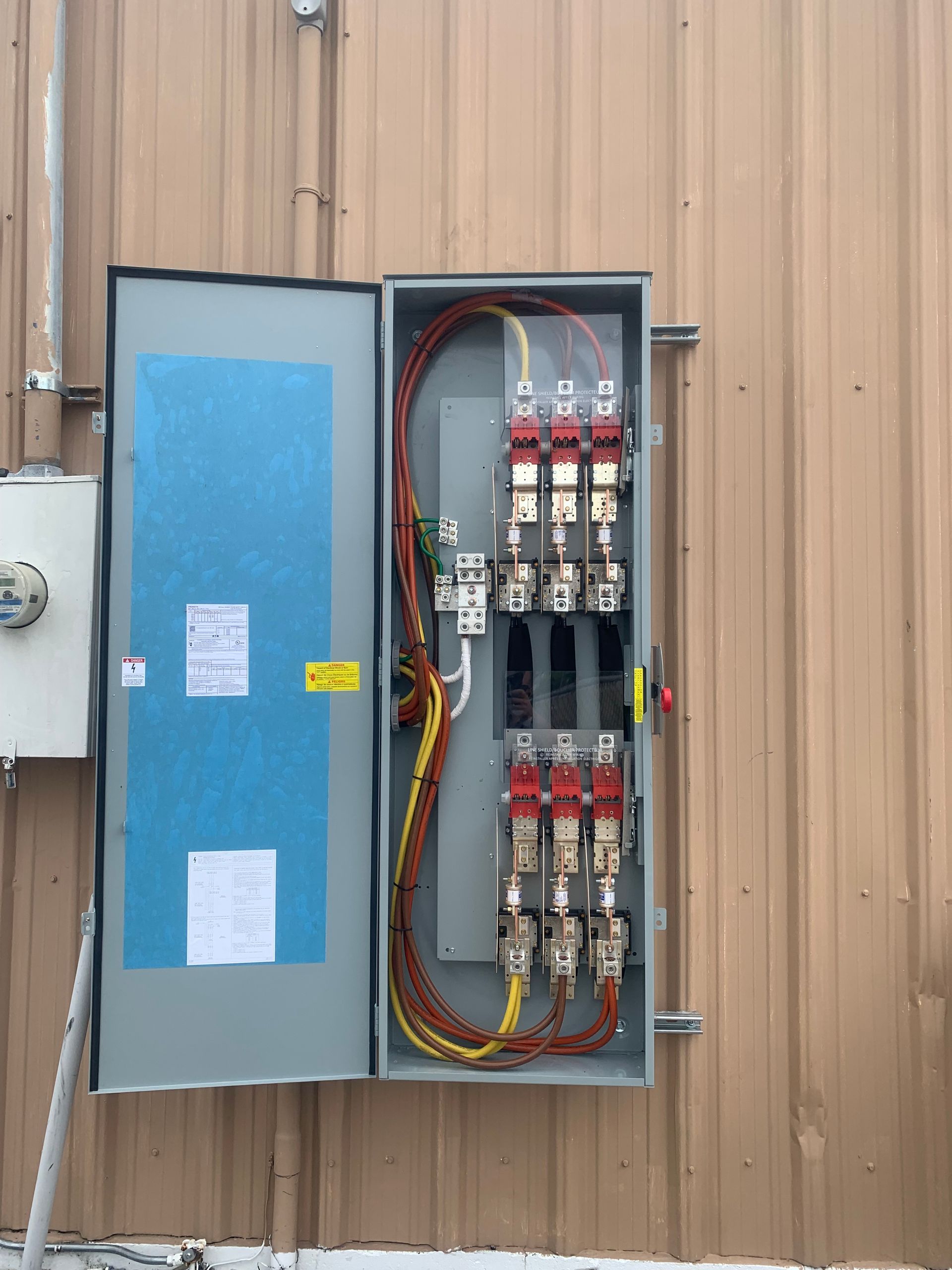 Open electrical breaker panel on tan metal wall with blue inner door and exposed wiring.