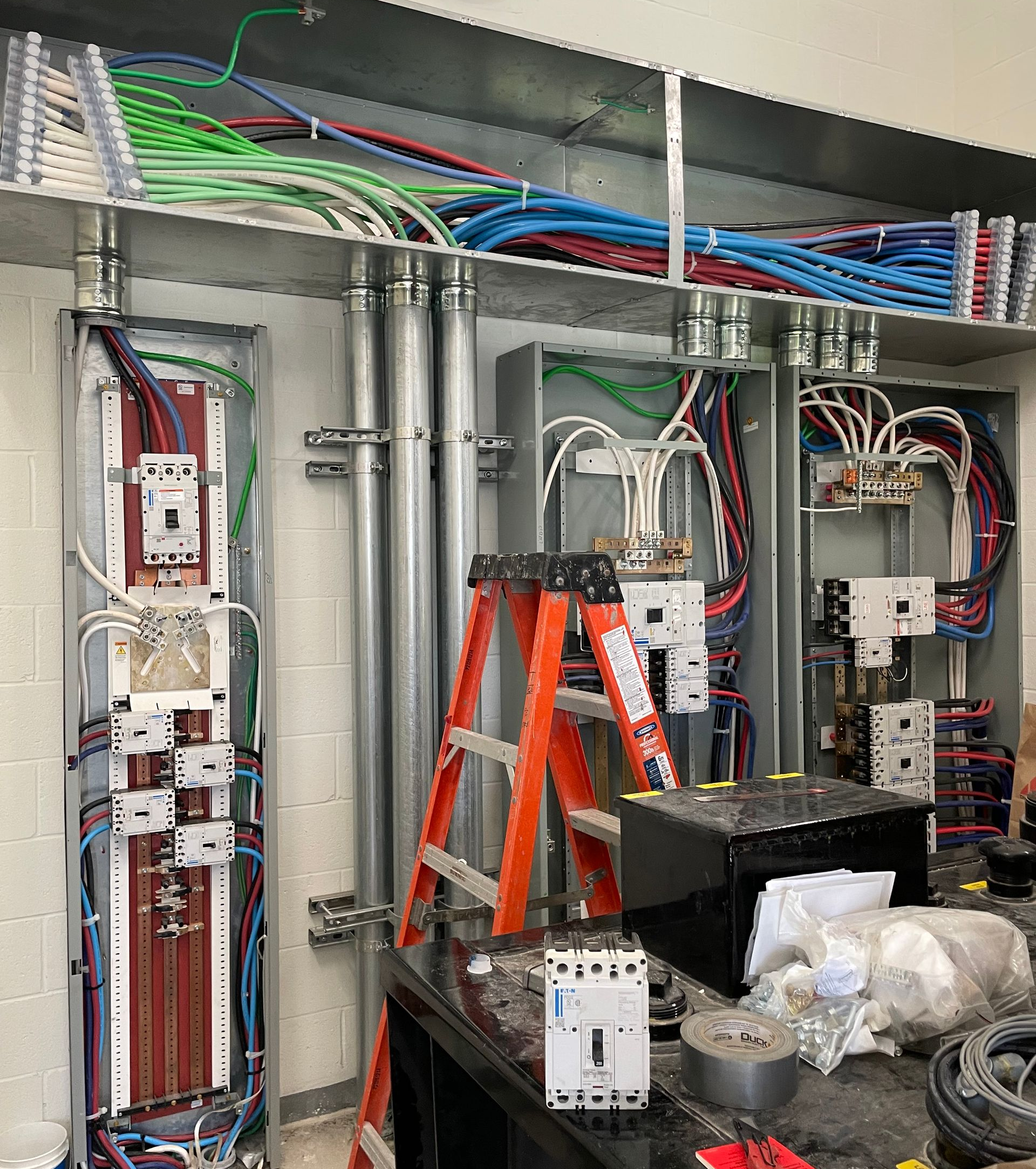 Electrical control cabinet interior with wiring, breakers, and orange ladder inside an industrial panel room