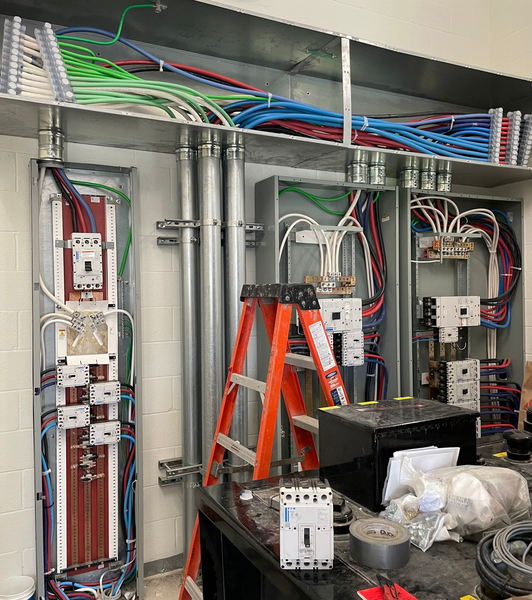 Electrical control cabinet interior with wiring, breakers, and orange ladder inside an industrial panel room
