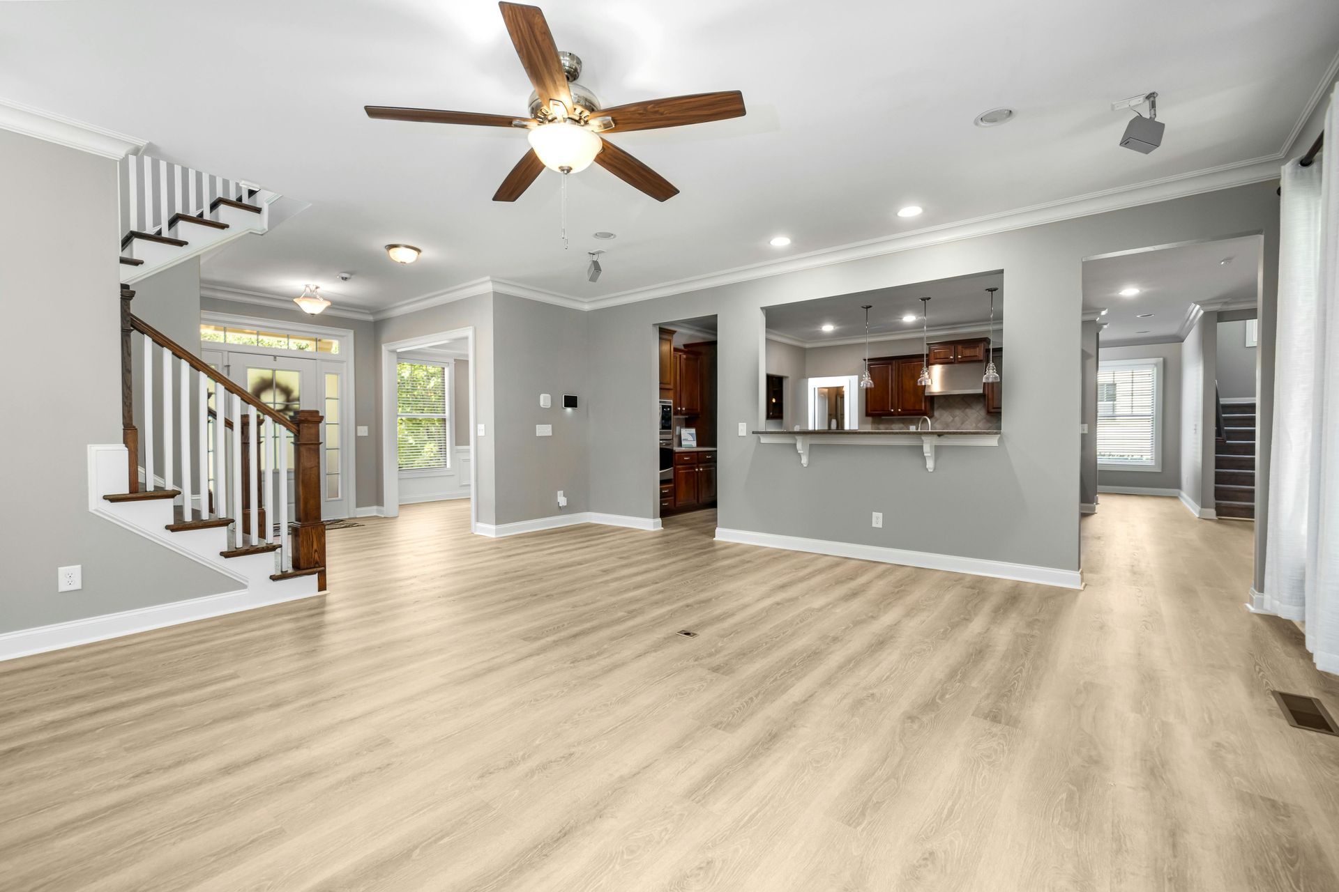 Bright open living room with light wood floors, gray walls, ceiling fan, stairs, and kitchen pass-through