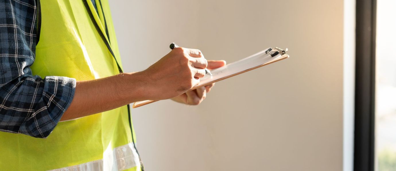 Person in a yellow safety vest holding a clipboard while writing indoors near a window