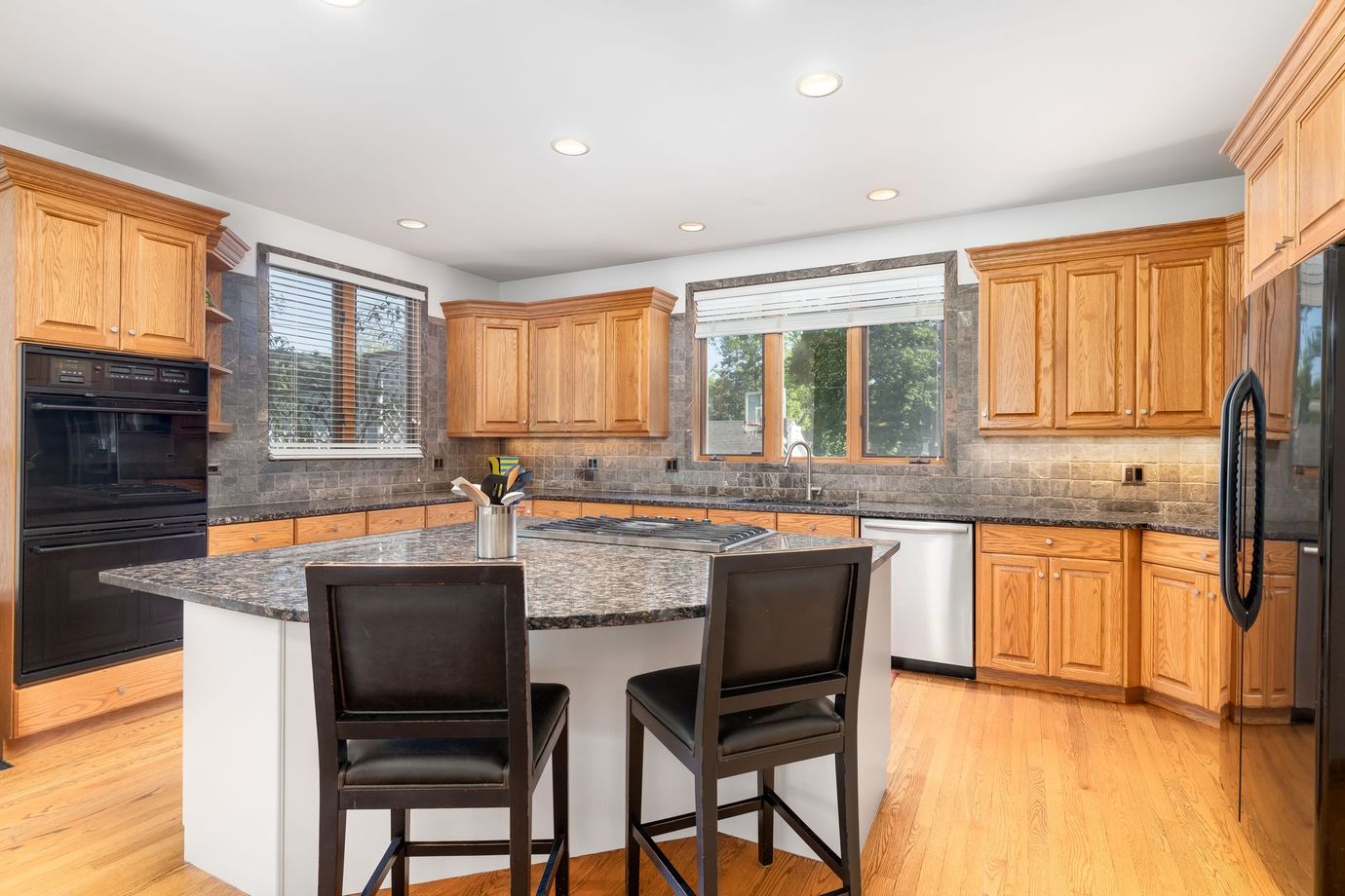 Bright kitchen with wood cabinets, granite island, black chairs, and stainless appliances