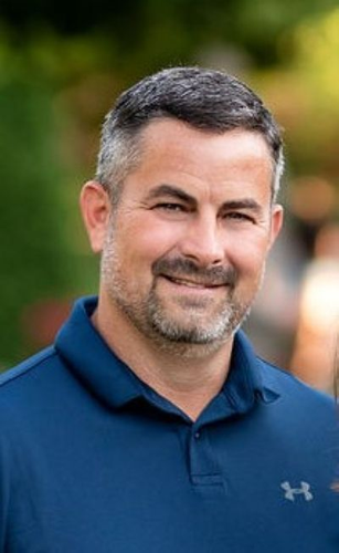 Smiling man in a blue polo shirt outdoors with blurred greenery in the background