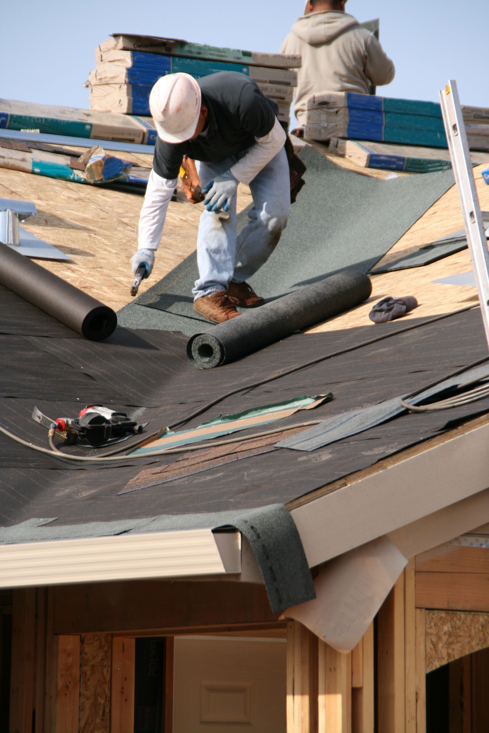 Workers installing roofing shingles on a house roof, with tools and rolled roofing materials nearby