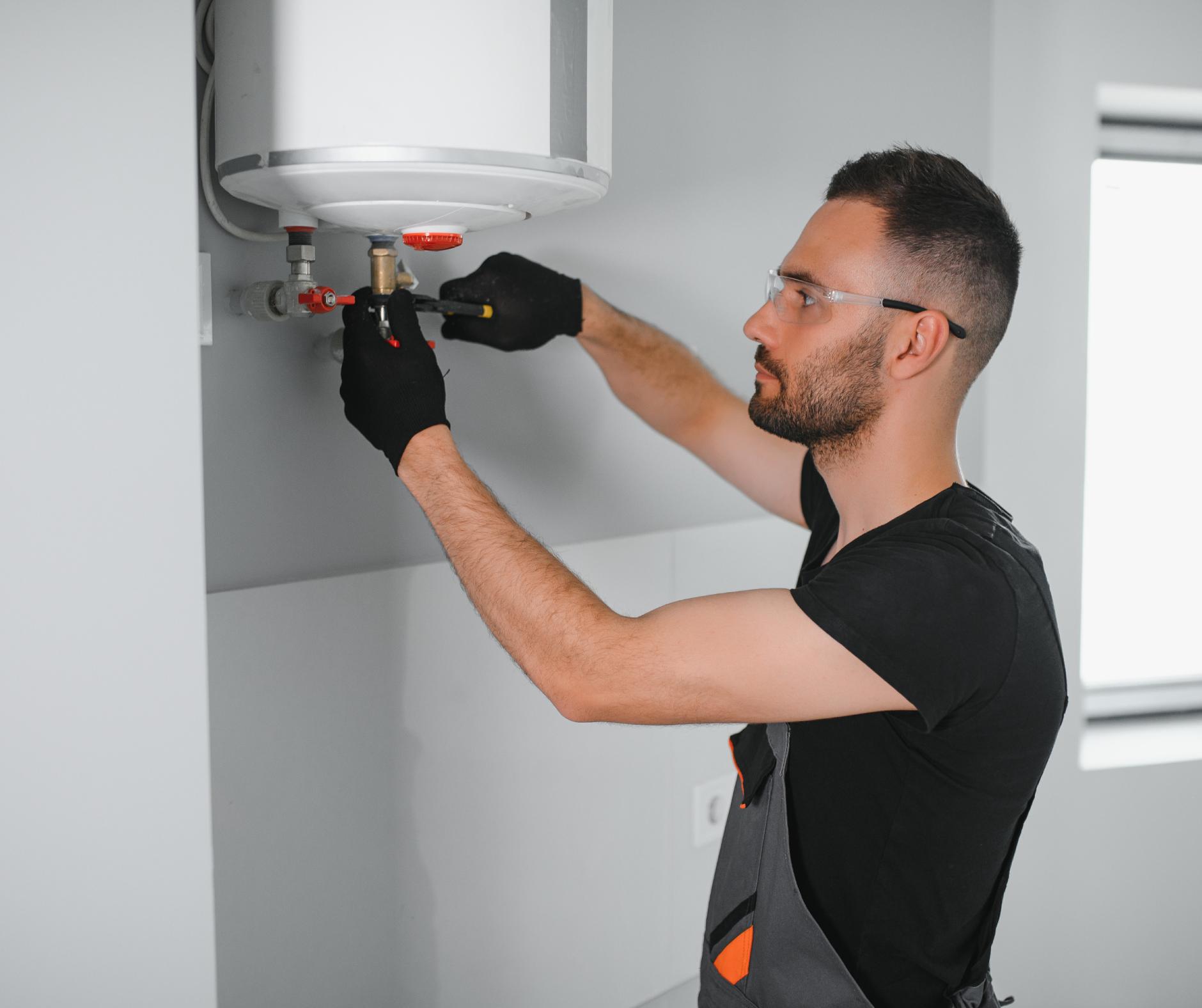 Man in gloves and safety glasses repairs water heater in a room.