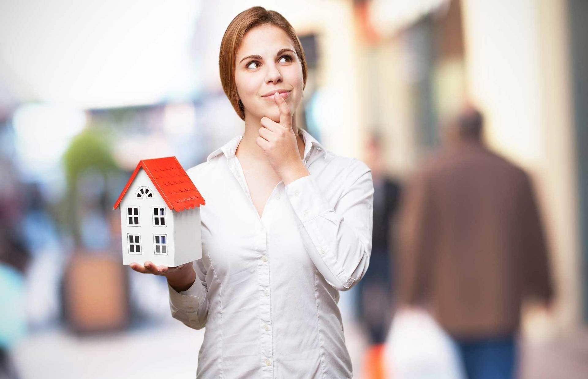 A person in a white shirt holds a small model house with a red roof, looking thoughtful with a hand to their chin.