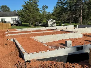 Construction site: Concrete block foundation framework, red dirt, blue sky, and a house in the background.