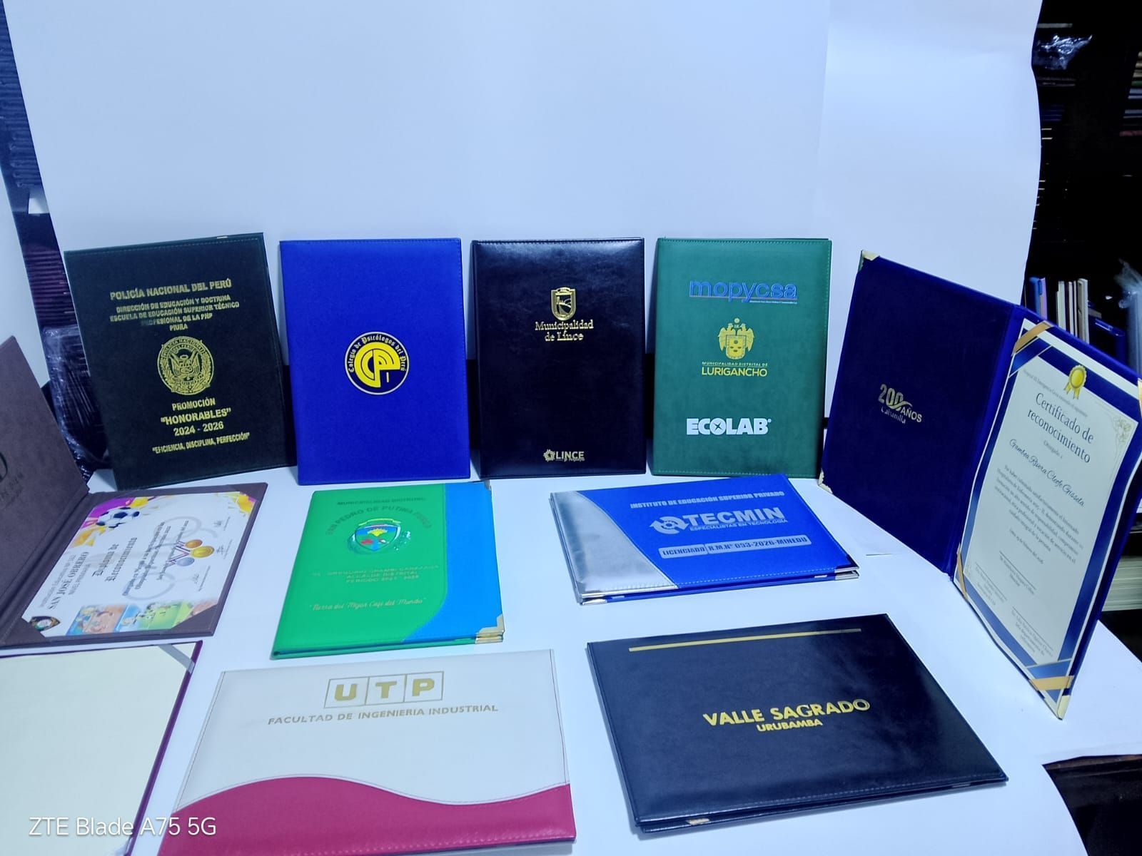 Varias carpetas azules, negras y verdes para diplomas o certificados, dispuestas sobre una superficie blanca.