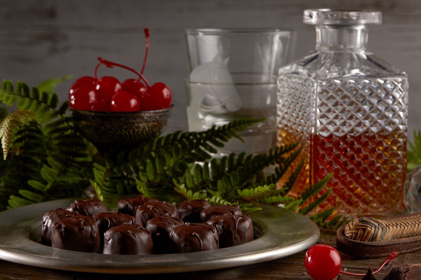 A plate of chocolate candies, cherries, and a whiskey decanter surrounded by ferns.