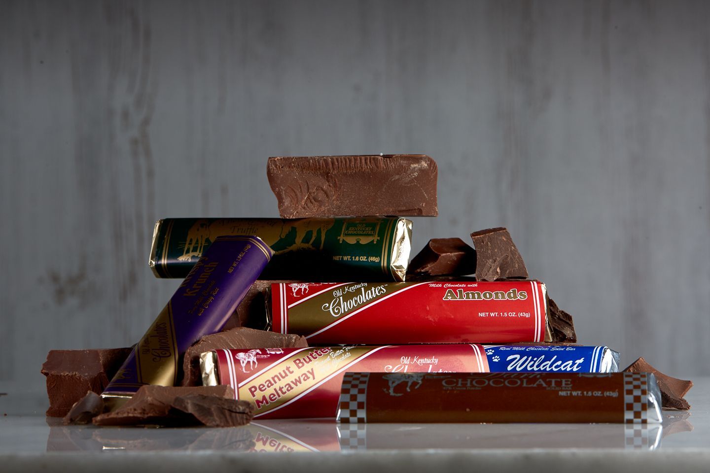 A stack of assorted chocolate candy bars and broken chocolate pieces on a white surface against a grey background.