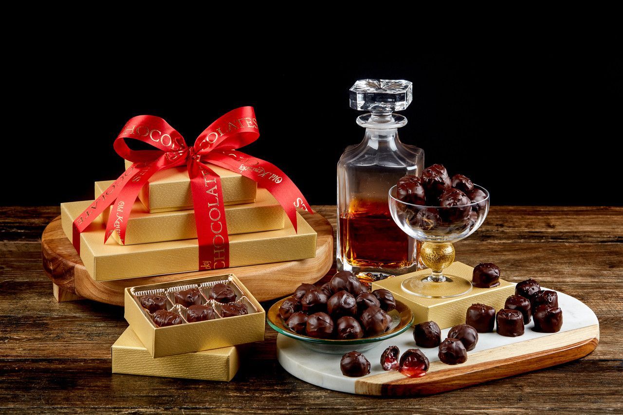 A stack of gift-wrapped chocolate boxes, a glass of dark chocolates, and a decanter on a rustic wooden table.