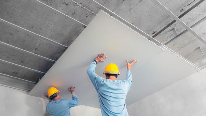 Two workers in yellow hard hats lifting a large white panel into place on a ceiling indoors