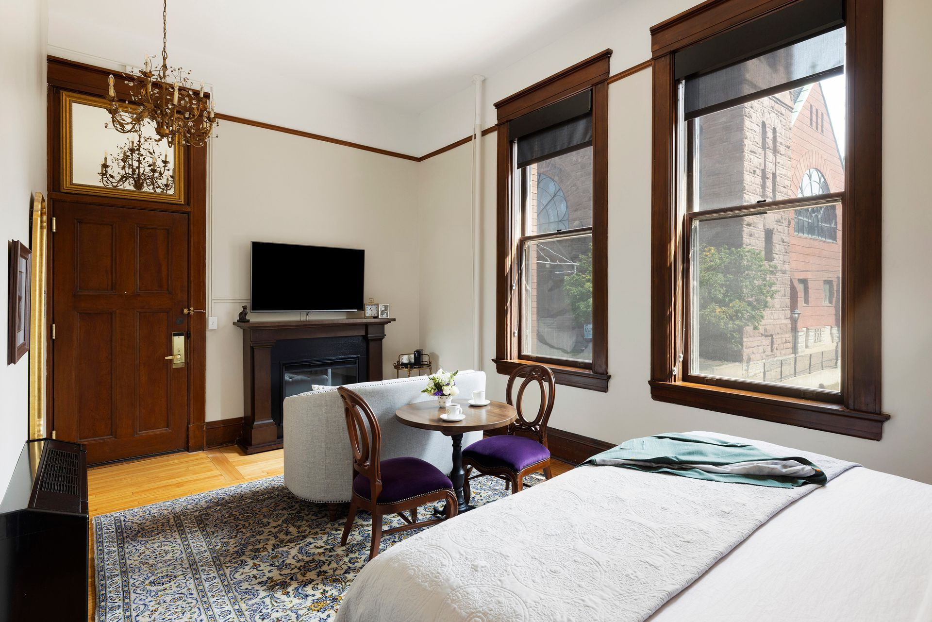 Bright bedroom with large windows, bed, desk, TV, and purple chair in a modern apartment room