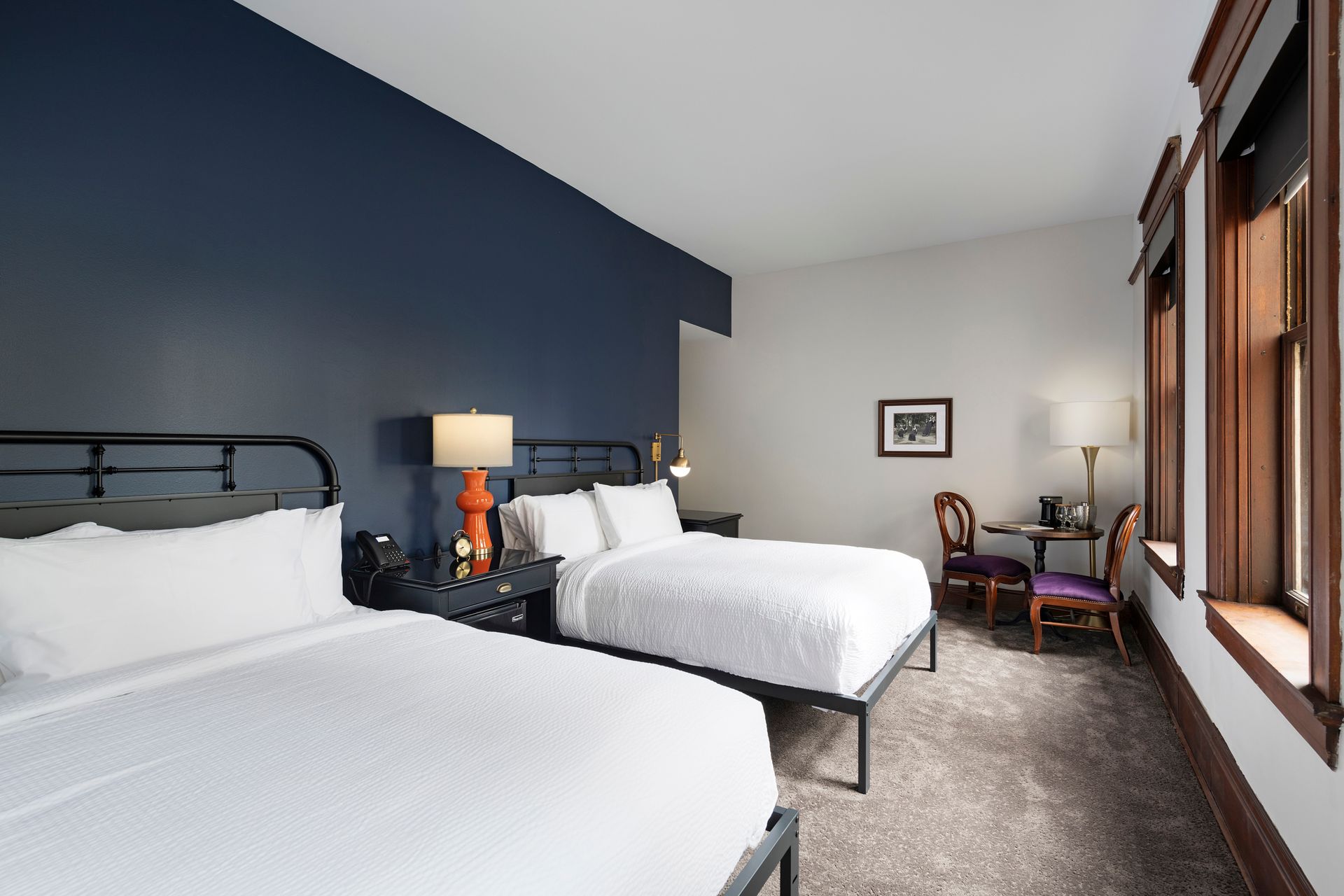 Hotel room with two white beds, dark blue accent wall, bedside lamp, and a windowed seating area