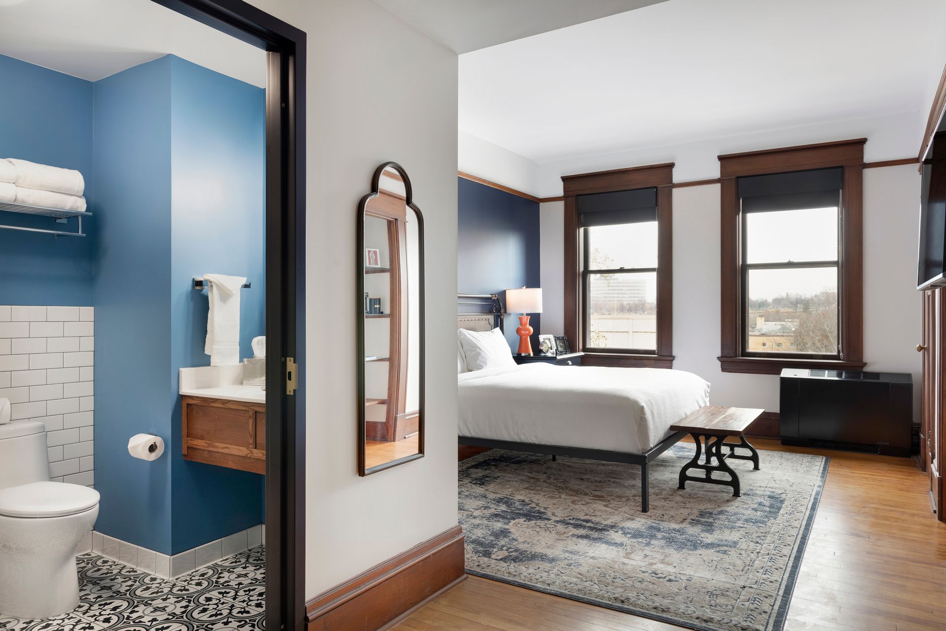 Modern bedroom with blue-accent bathroom doorway, white bedding, wood floors, and large windows.