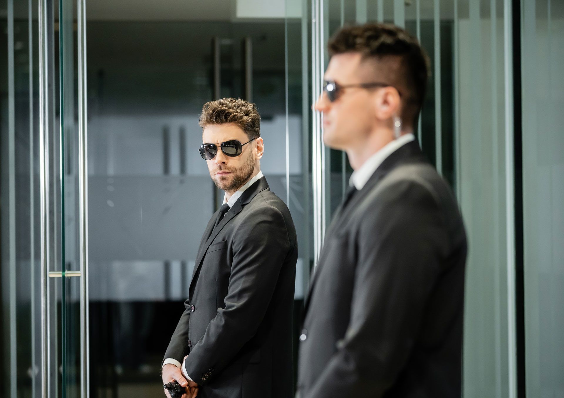 Two men in dark suits and sunglasses standing in a glass office corridor.